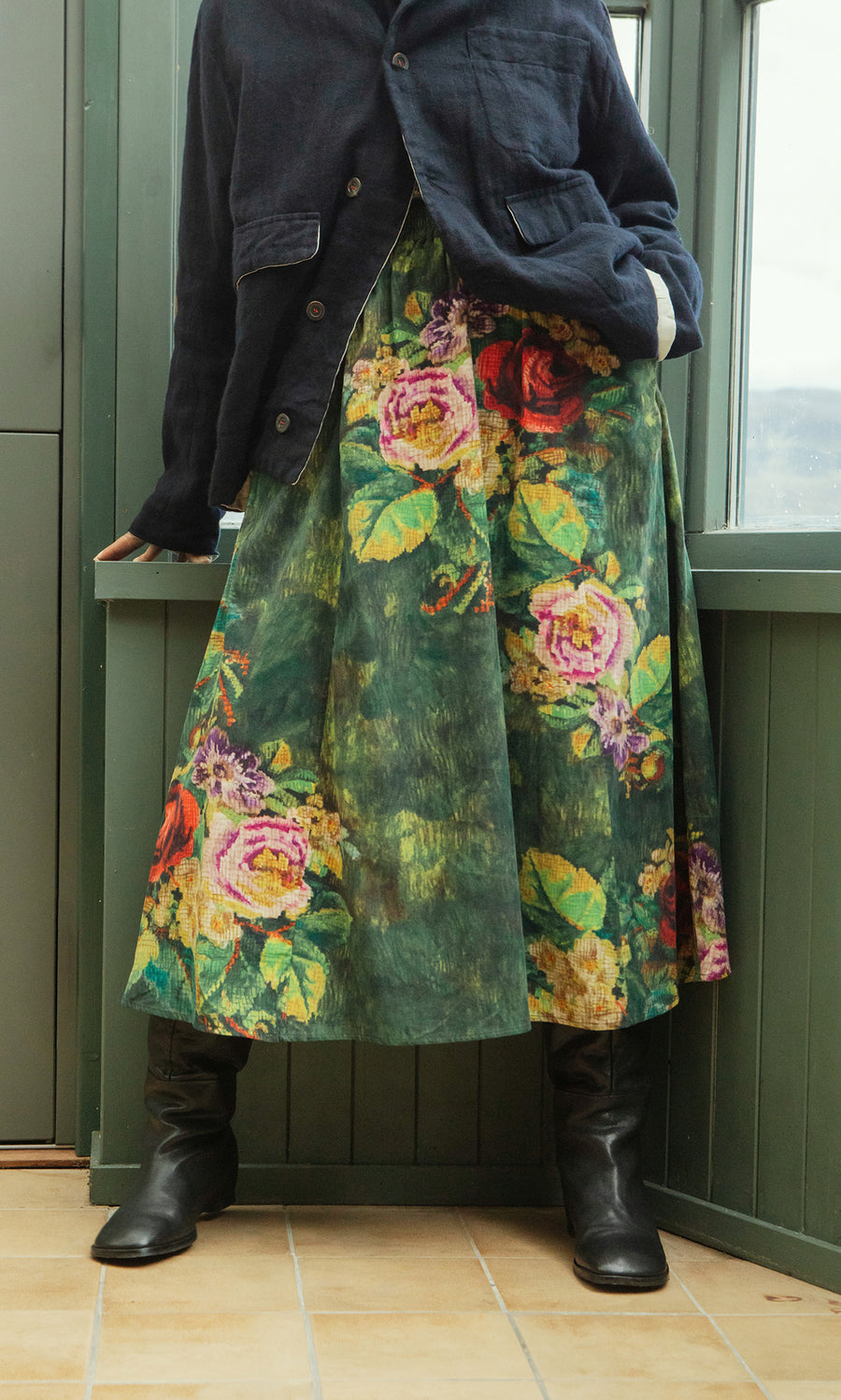 Plümo  Person in a dark jacket and Verity Skirt stands indoors by a window, wearing black boots.