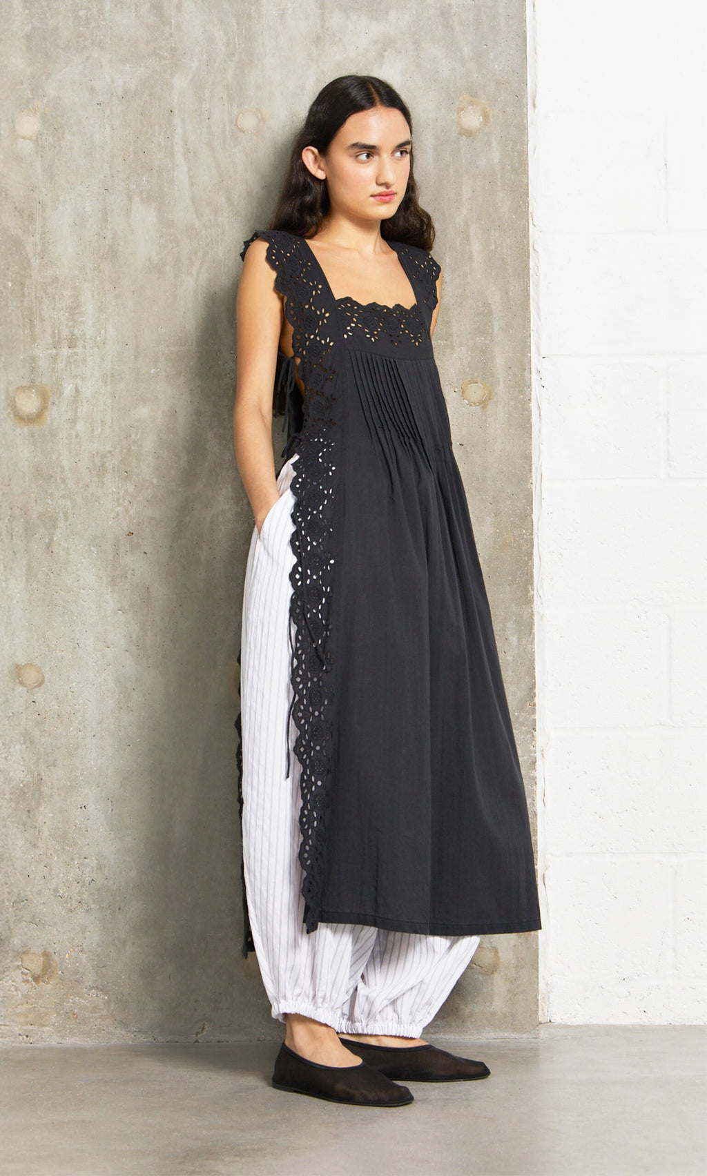 Plümo- Woman leans on a concrete wall wearing the Vintage Lace Dress over white striped pants and black flats.