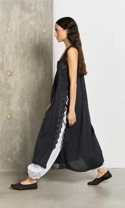 Plümo- Woman in a Vintage Lace Dress over white pants walks by a concrete and white brick wall indoors.