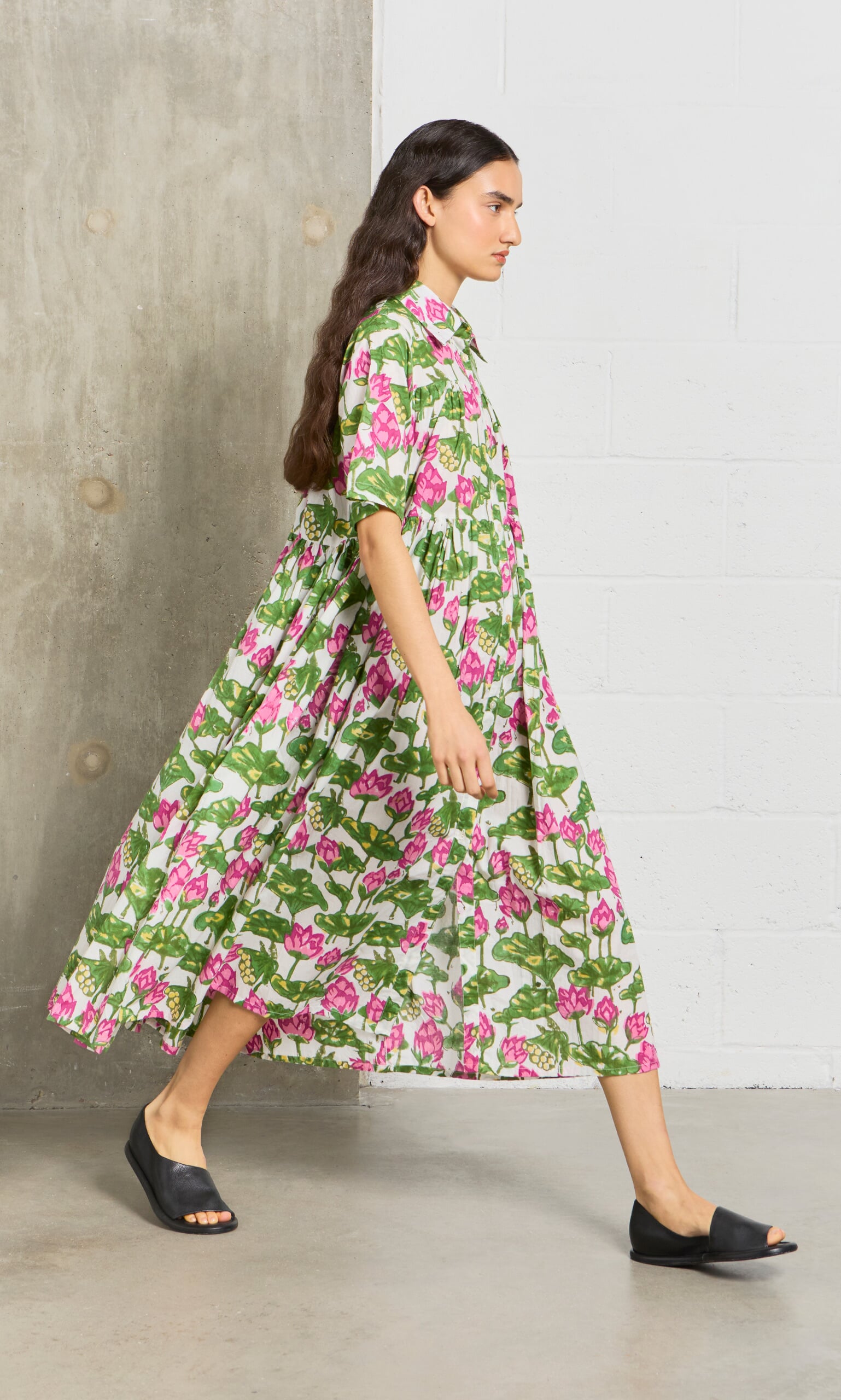 Plümo- A woman in the Waterlily Dress, featuring block-printed cotton voile, walks in black slides by a concrete wall.
