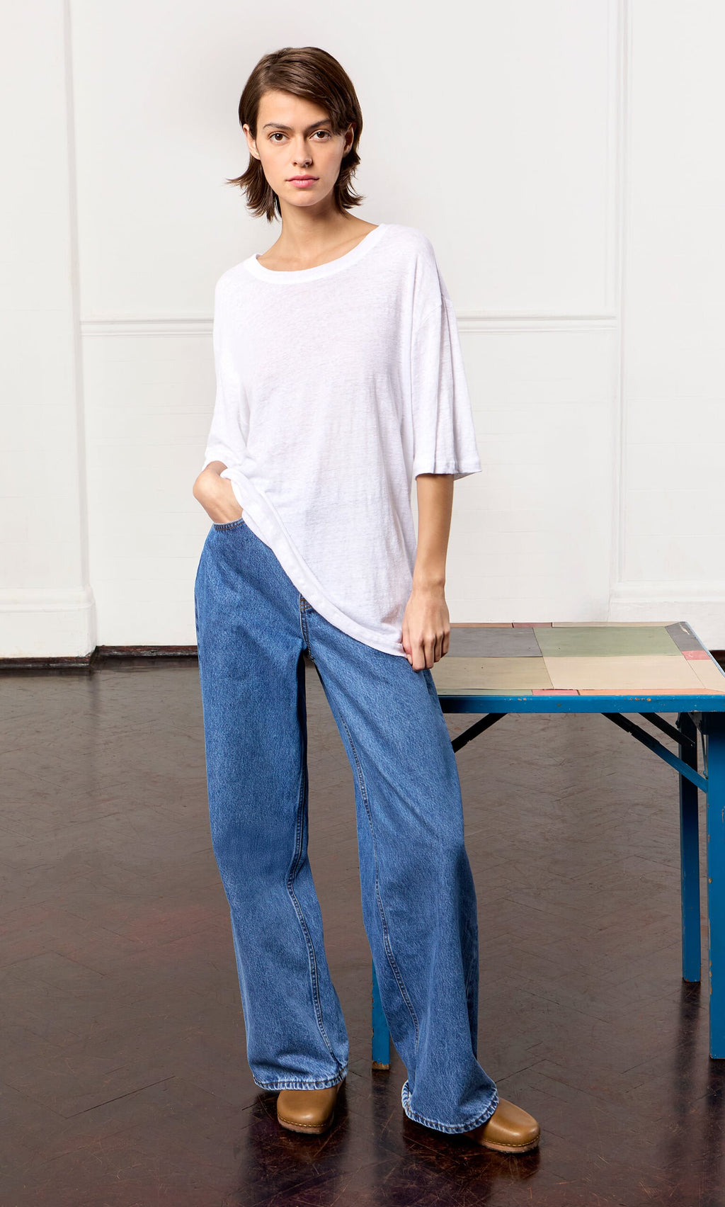 Plümo- Someone in a relaxed fit White Tee, blue wide-leg jeans, and brown shoes stands indoors beside a table.