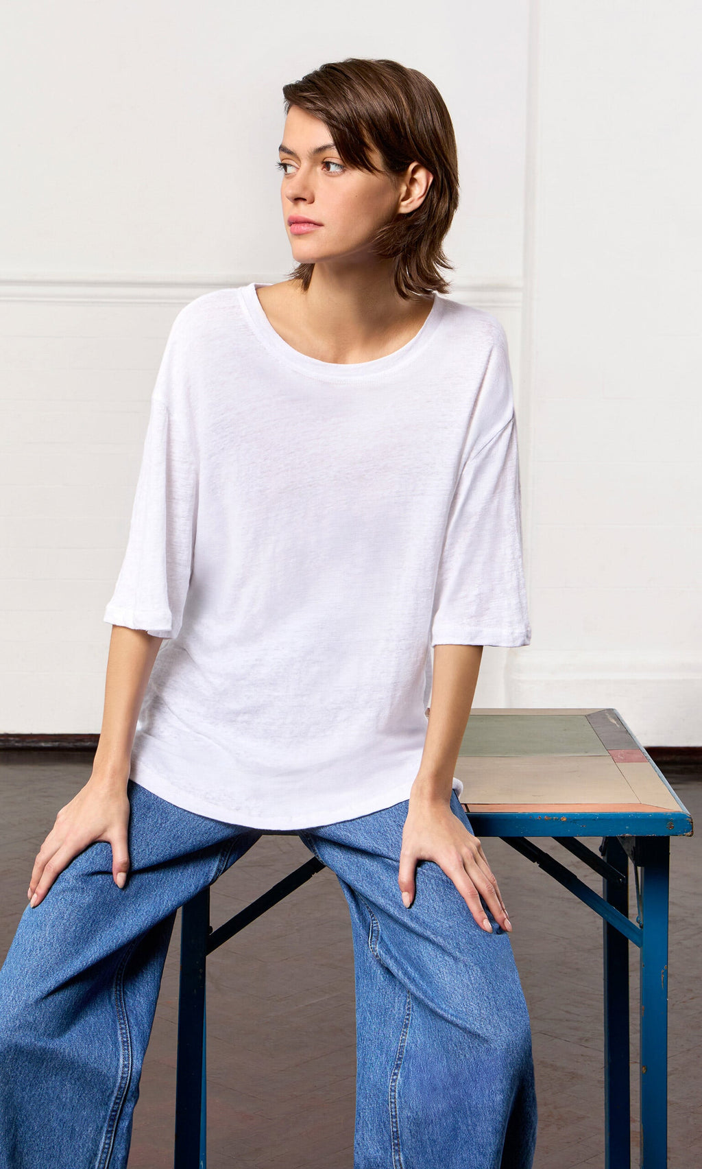 Plümo- Wearing a relaxed fit White Tee and blue jeans, a person sits on a table in a bright room, looking to the side.