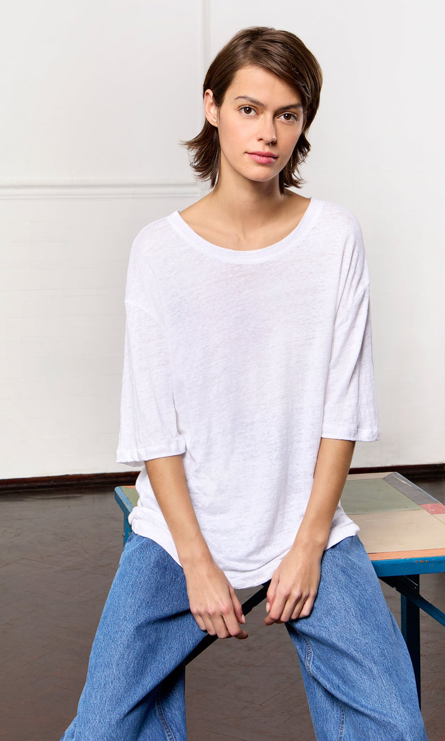 Plümo- Person in a relaxed fit White Tee and blue jeans sits on a table, looking at the camera indoors.