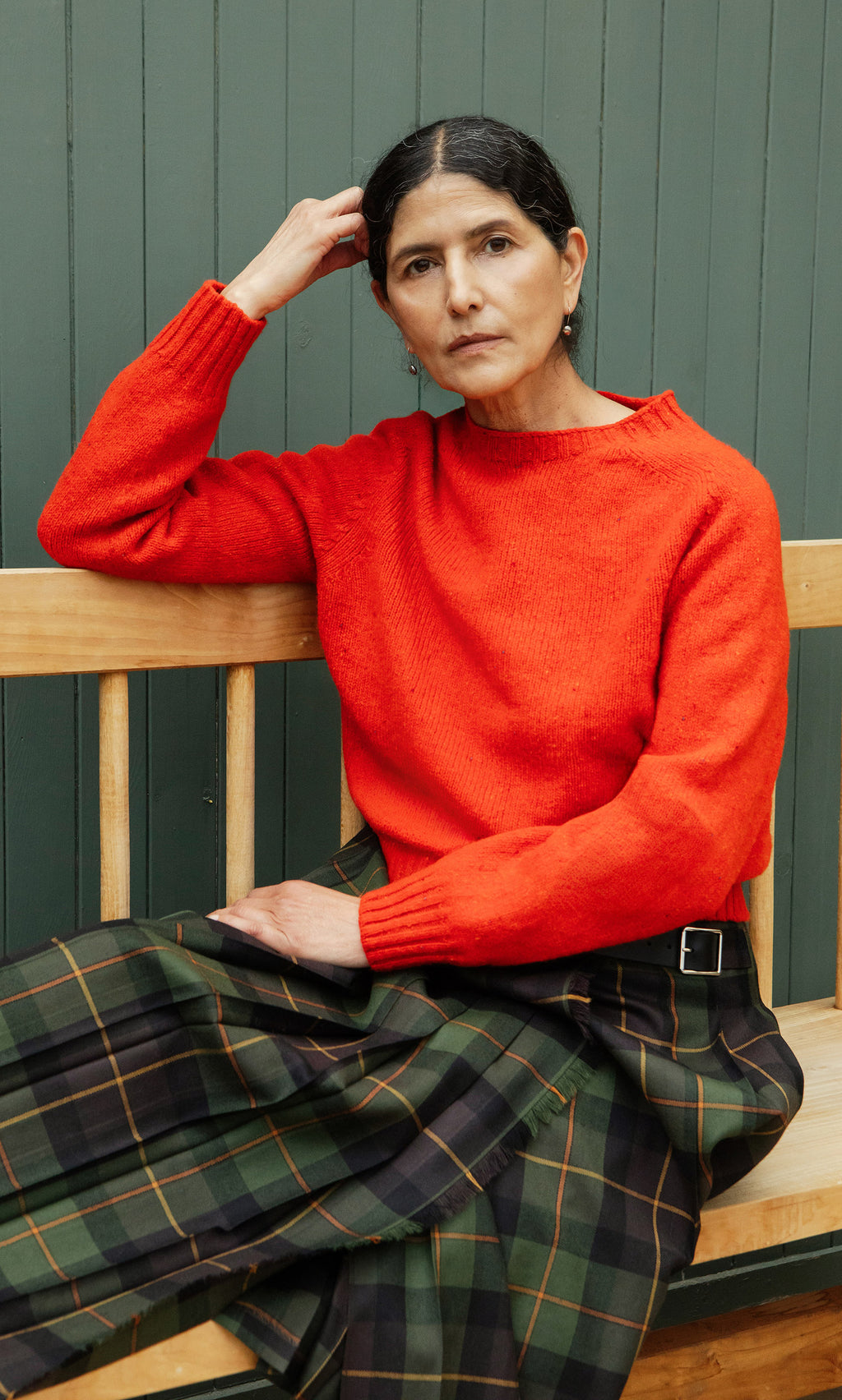 Plümo  Woman in the Wicklow Crewneck - Red Fleck sits on a bench, gazing ahead against a green paneled wall.
