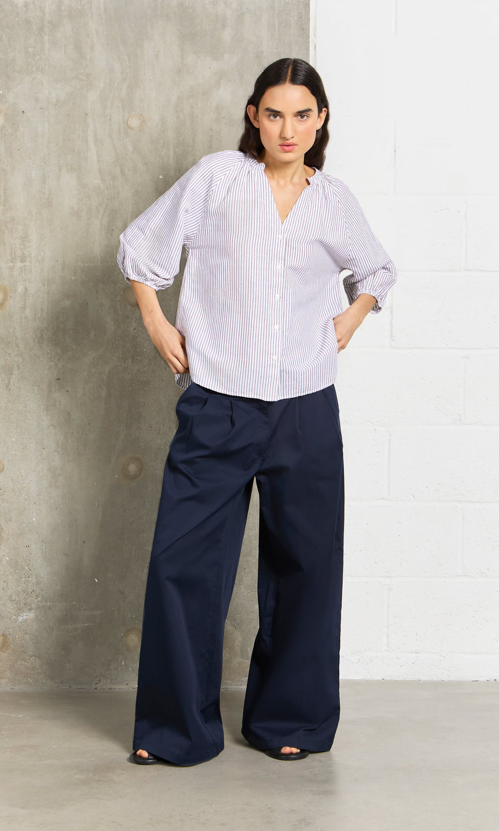 Plümo- Woman in wide navy chinos and a striped button-up stands by a concrete and white wall.