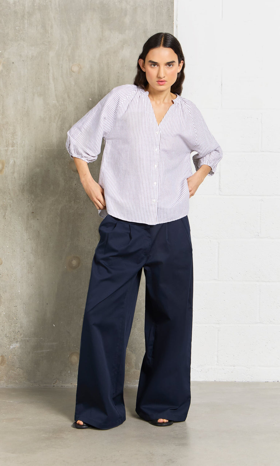 Plümo- Woman in wide navy chinos and a striped button-up stands by a concrete and white wall.