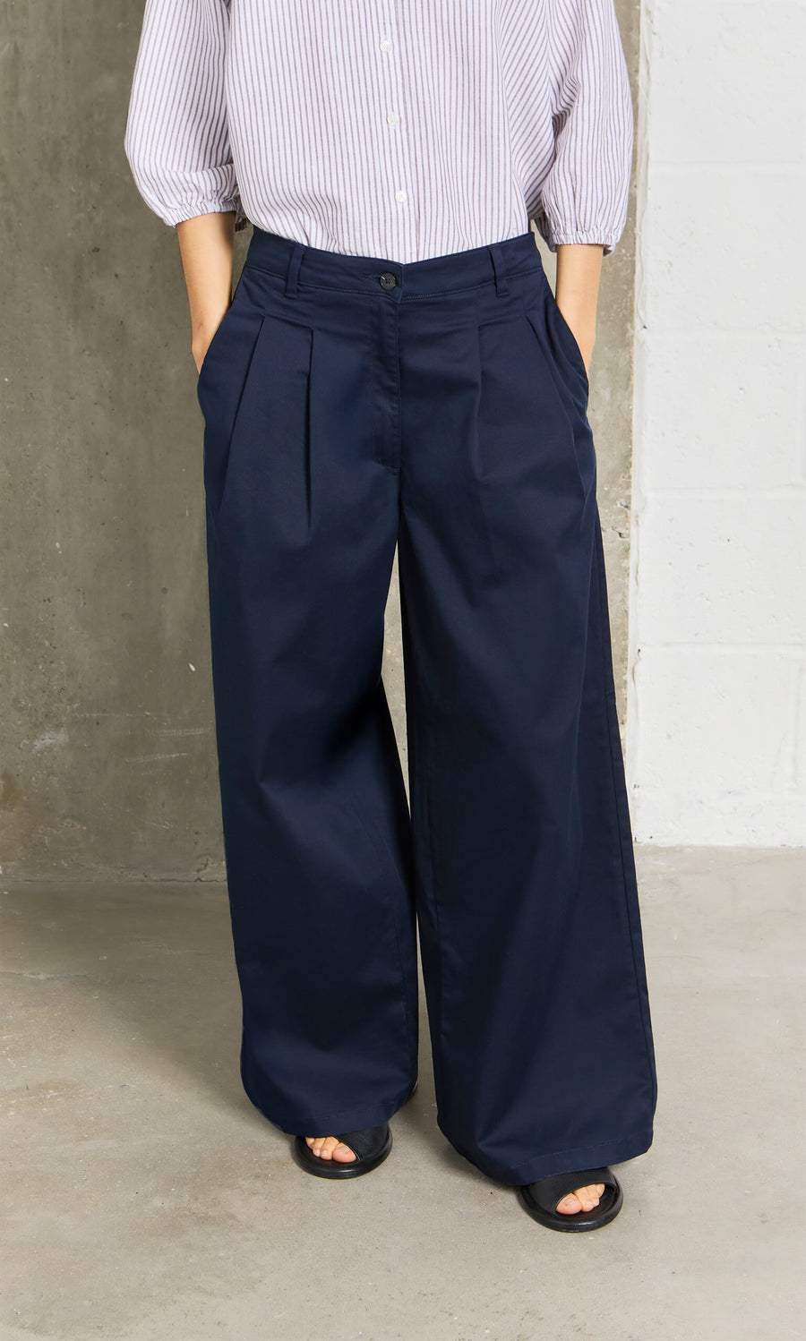 Plümo- Person wearing Wide Navy Chinos, black sandals, and a striped shirt, standing by a concrete and white wall.