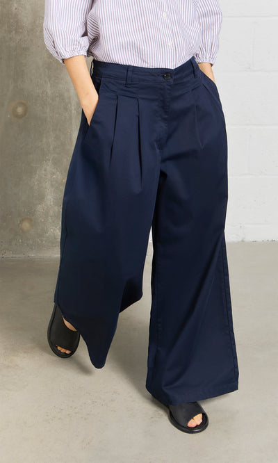Plümo- Person in Wide Navy Chinos, black sandals, and a striped shirt standing by a brick wall.