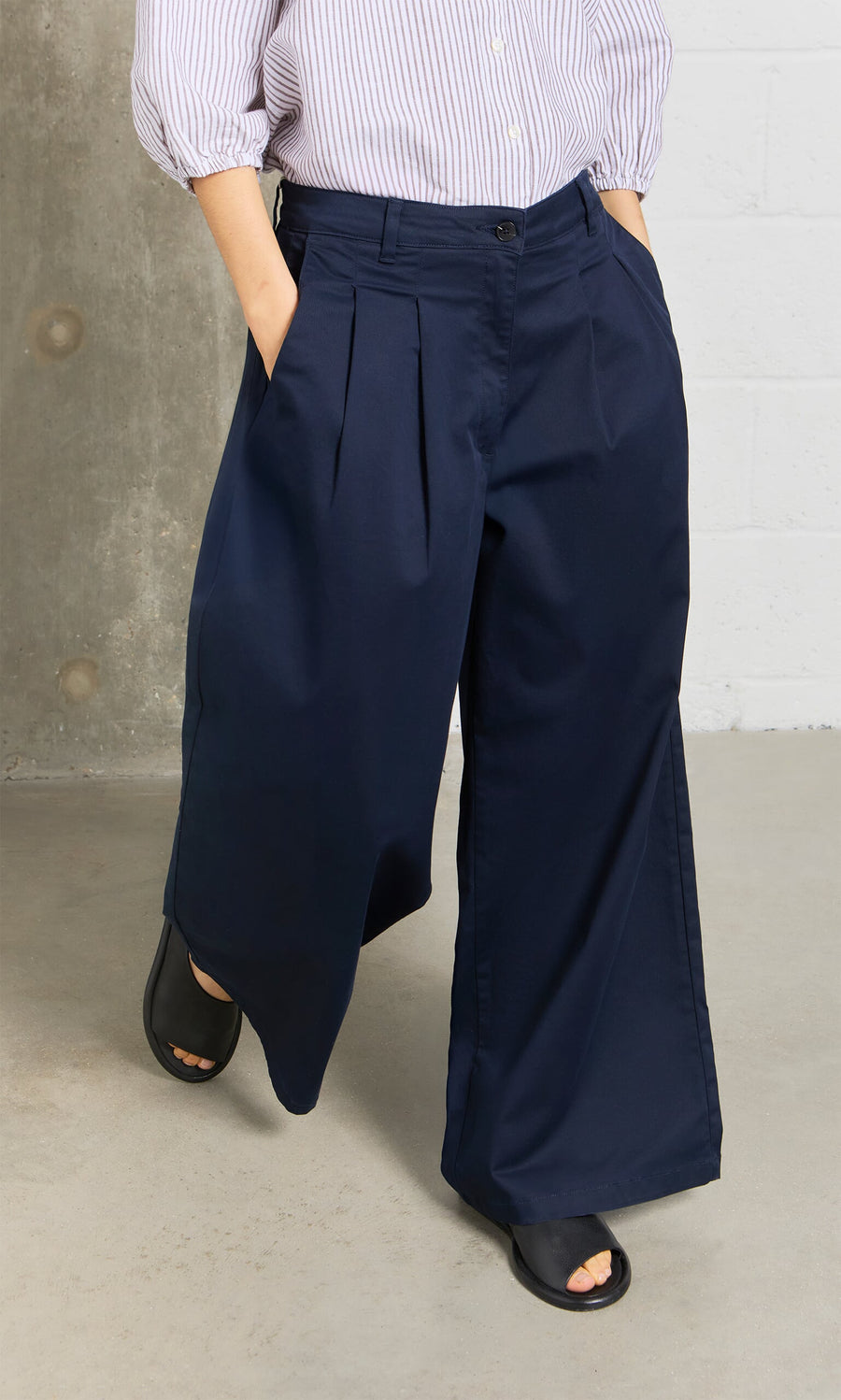 Plümo- Person in Wide Navy Chinos, black sandals, and a striped shirt standing by a brick wall.