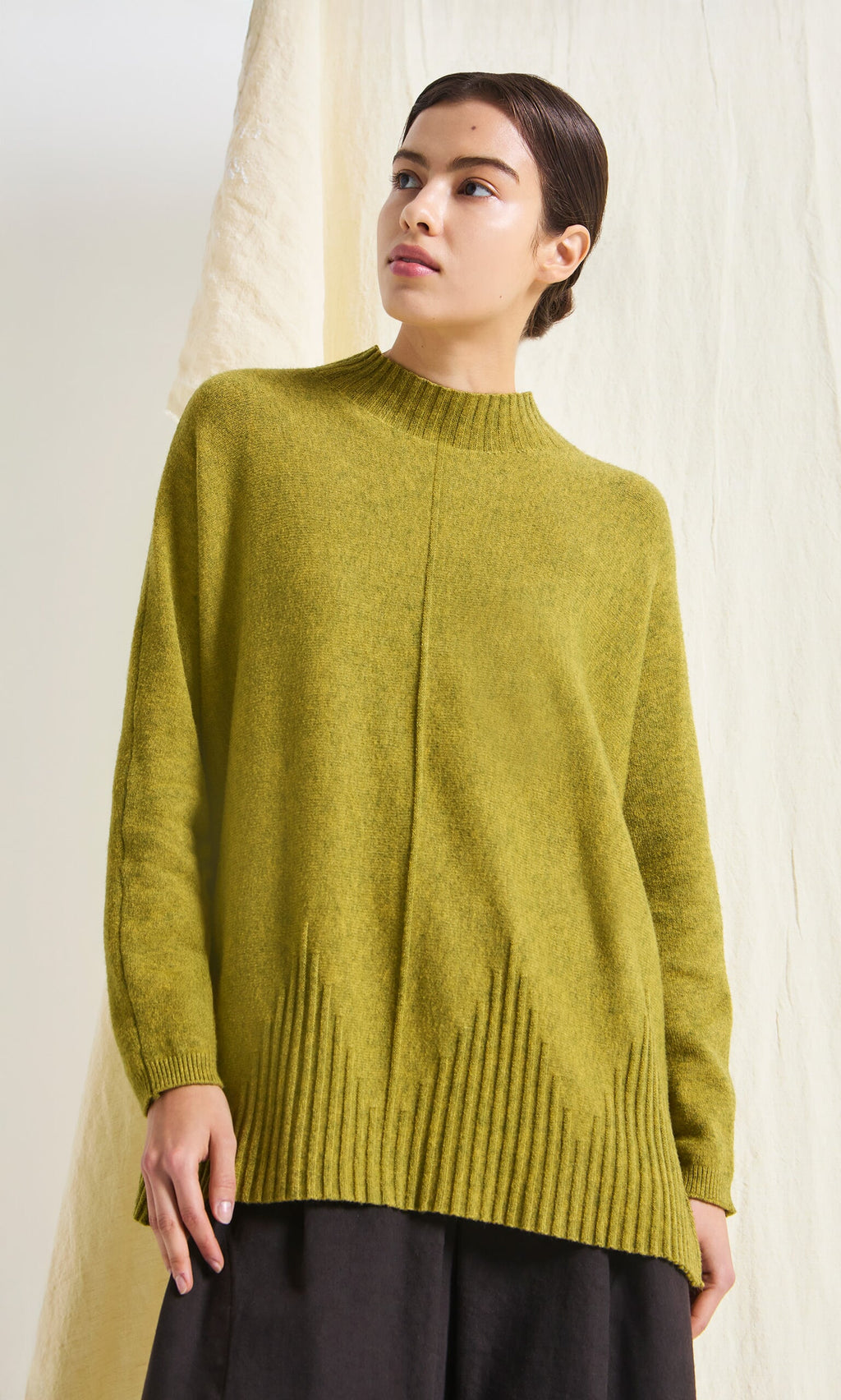 Plümo- Woman wearing the Wild Millet Jumper in green stands before a light, textured background, looking slightly to the side.