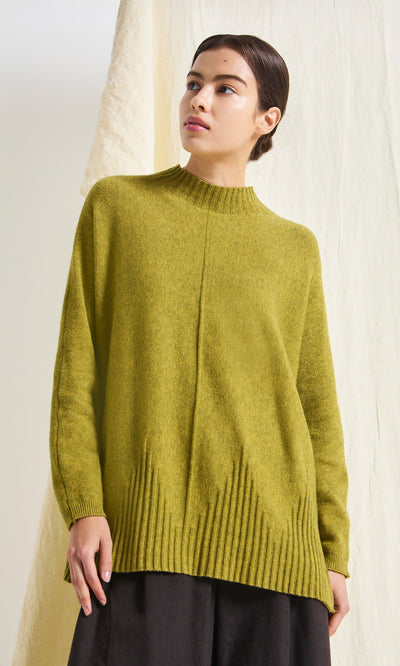 Plümo- Woman wearing the Wild Millet Jumper in green stands before a light, textured background, looking slightly to the side.