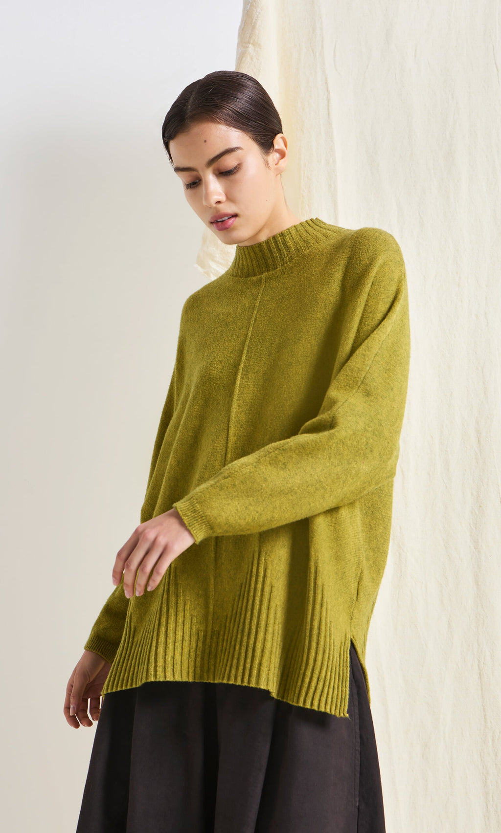 Plümo- Woman in a marled Wild Millet Jumper and black skirt, looking down with a neutral expression.