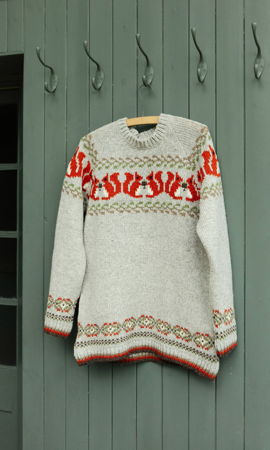 Plümo  The Woodland Jumper, a chunky knit with red and green patterns, hangs on a hanger against a green paneled wall.