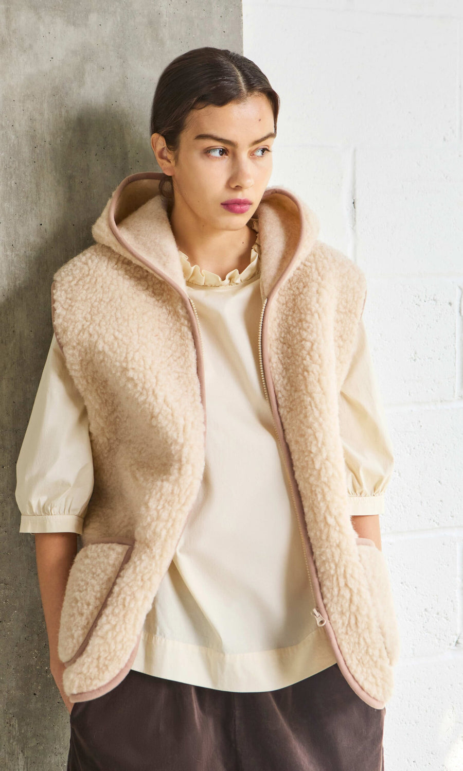 Plümo- Woman wearing the Wool Gilet - Natural over a light blouse, standing by a concrete and white brick wall.