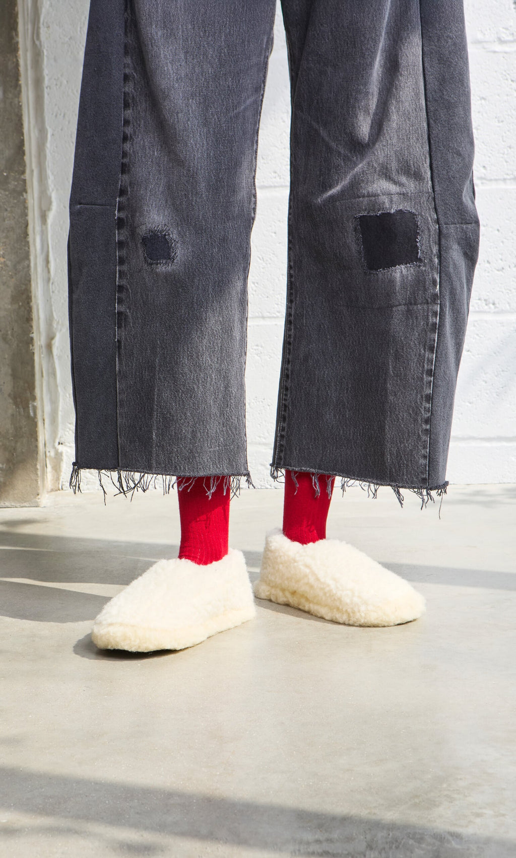 Plümo- Person in frayed black jeans, red socks, and Wool Slippers - Natural stands on a concrete floor.