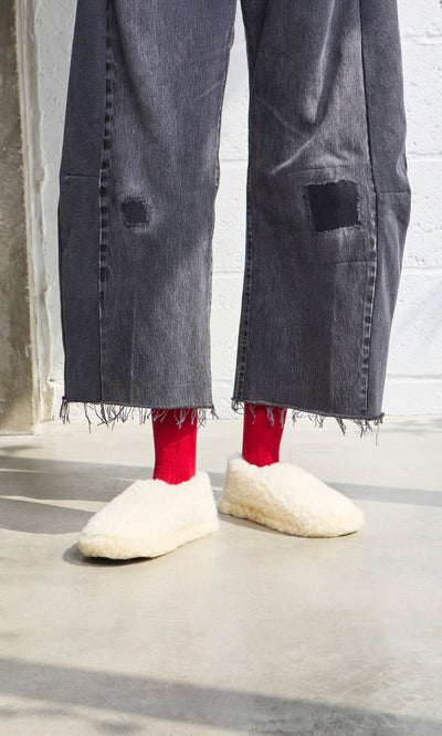Plümo- Person in frayed black jeans, red socks, and Wool Slippers - Natural stands on a concrete floor.