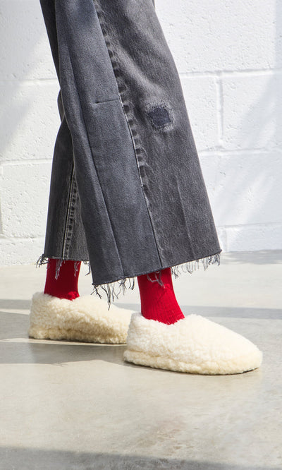 Plümo- Person wearing frayed black jeans, bright red socks, and Wool Slippers - Natural with a non-slip sole on concrete floor.
