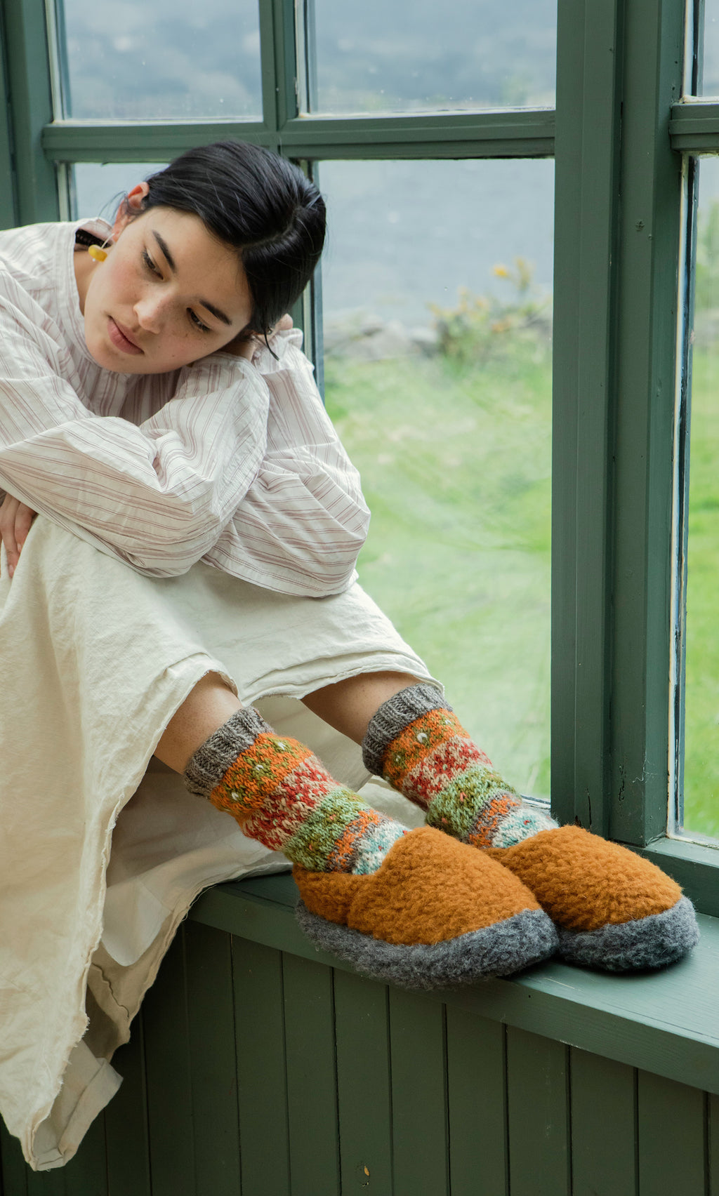 Plümo  A woman in comfy clothes and Wool Slippers - Caramel sits by a window, gazing down thoughtfully.