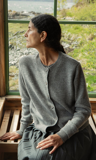 Plümo  Woman in a Grey Melange Cardigan sits by a window on a wooden bench, gazing outside at nature.