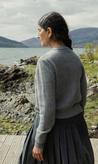 Plümo  Woman with dark braids wears a Grey Melange Cardigan by a rocky lakeshore, mountains in the background.