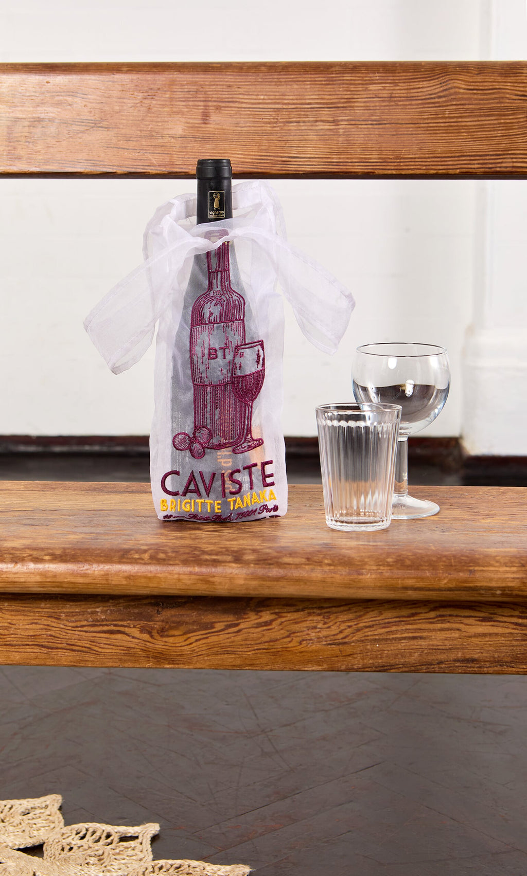 Plümo- A Caviste Wine Bottle Gift Bag, a wine bottle, glass, and tumbler sit on a wooden bench against a white wall.
