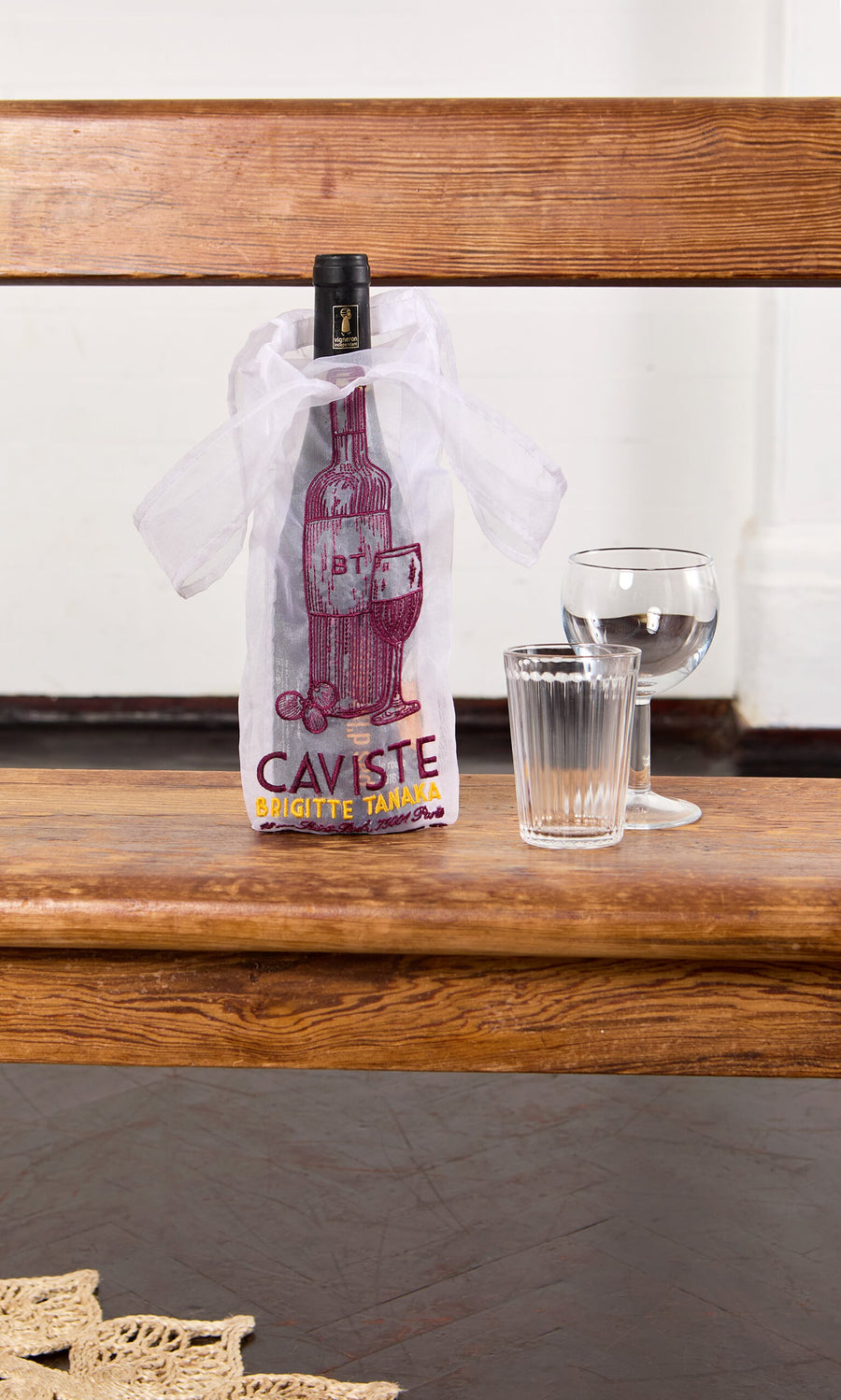 Plümo- A Caviste Wine Bottle Gift Bag, a wine bottle, glass, and tumbler sit on a wooden bench against a white wall.
