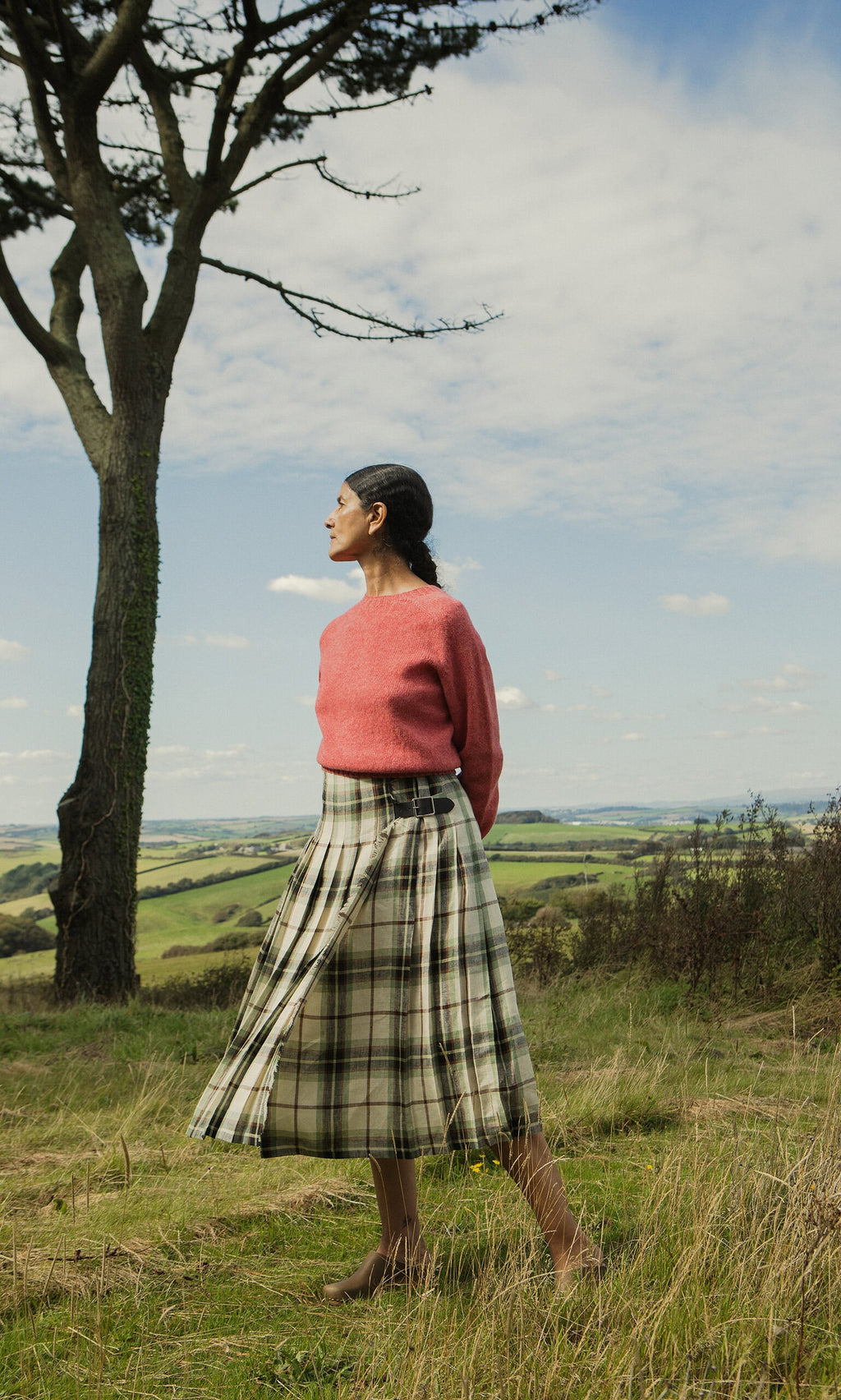Plümo- Woman wears Wicklow Crewneck - Tea Rose, standing in a grassy field with a tree and rolling hills behind.