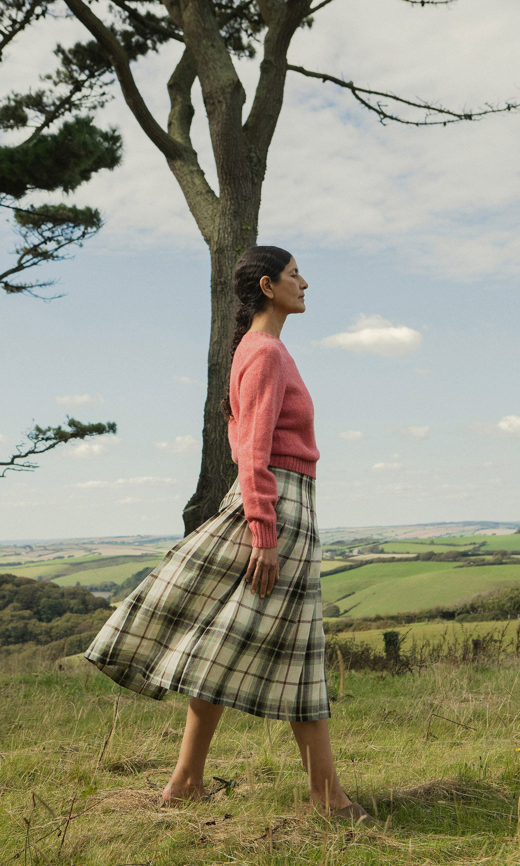 Plümo- Woman in pink sweater and Hawksmoor Kilt walks in grassy field with rolling Dublin hills in the background.