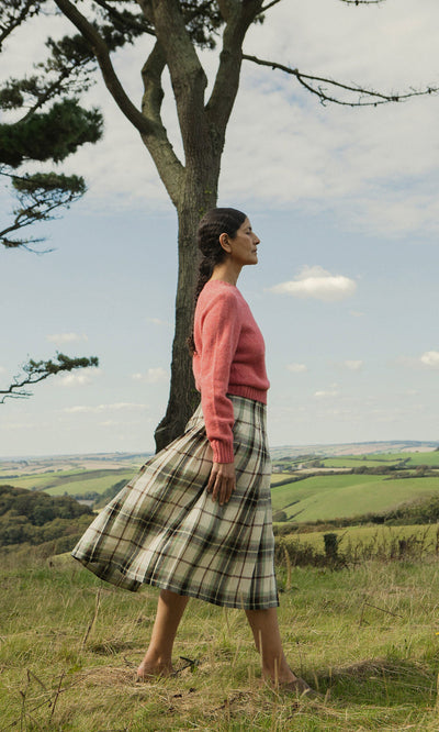 Plümo- Woman in pink sweater and Hawksmoor Kilt walks in grassy field with rolling Dublin hills in the background.