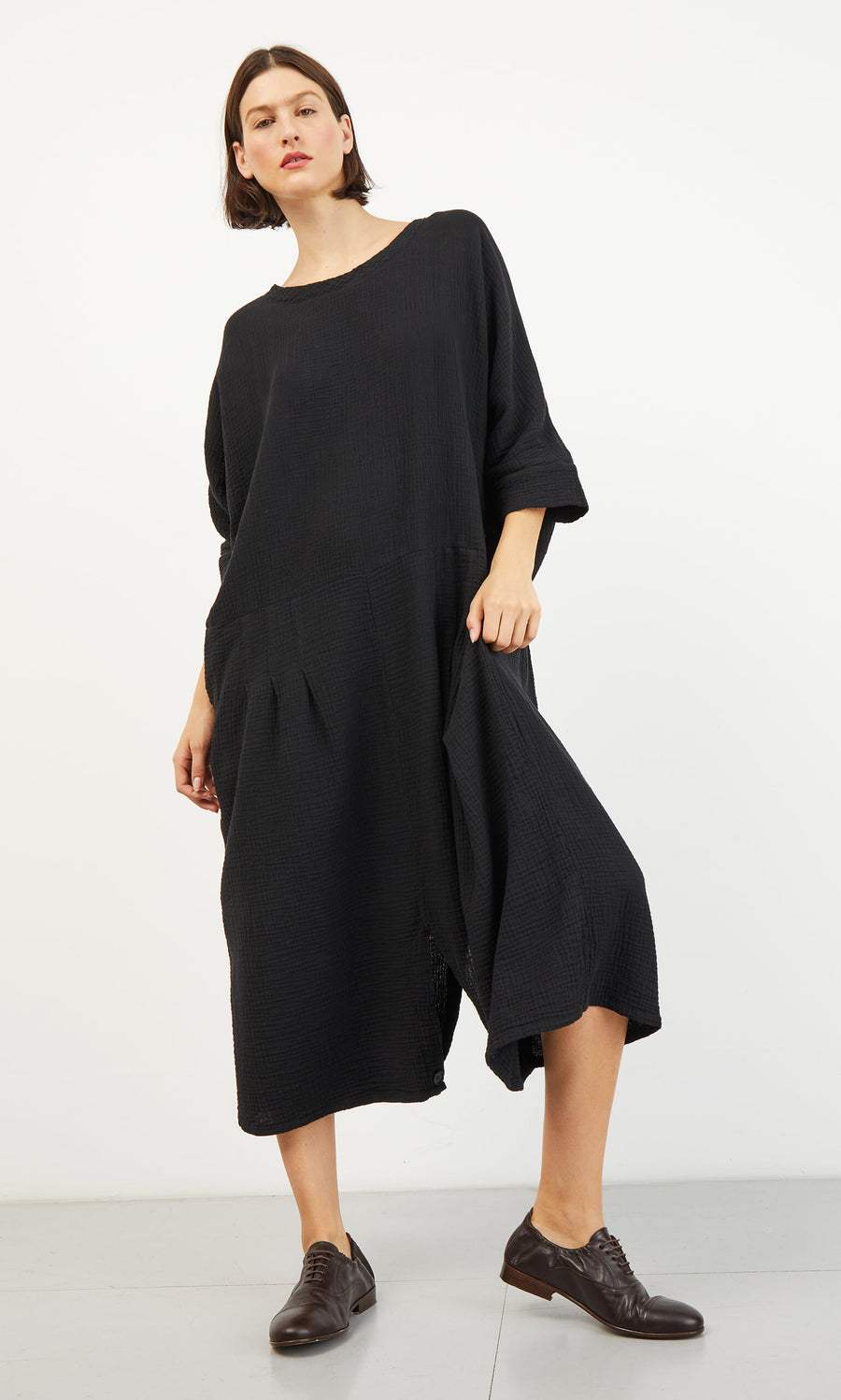 Plümo- Woman in a loose-fitting black Culotte Dress and brown oxford shoes stands against a plain white background.