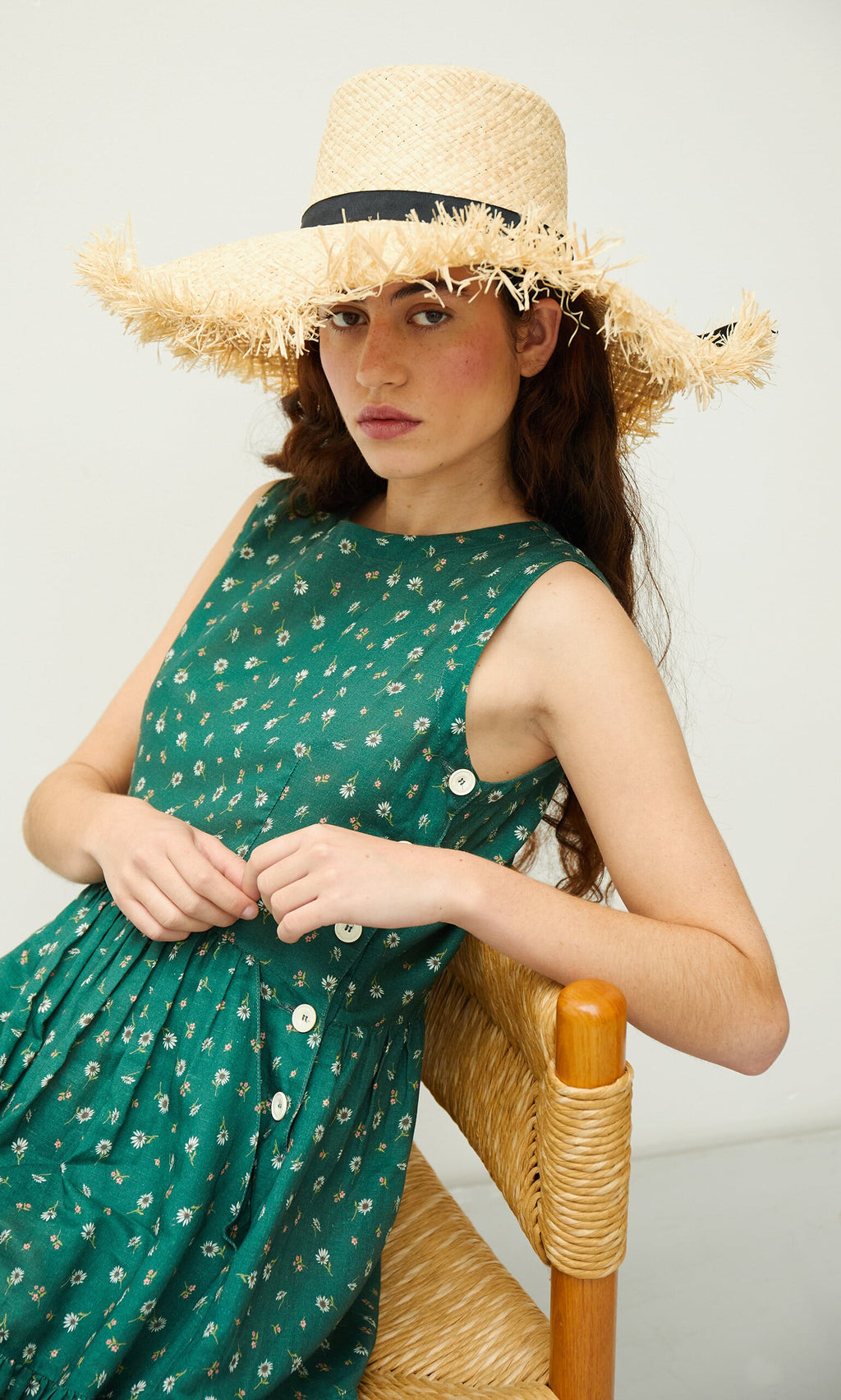Plümo- Woman in a green floral dress and Frayed Straw Hat sits on a woven chair, looking at the camera.