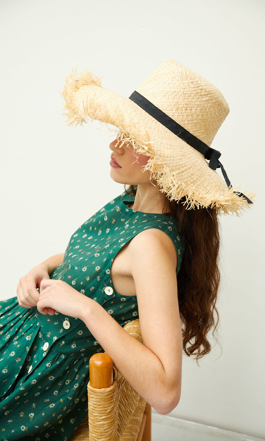 Plümo- Woman in a green patterned dress sits sideways, wearing the Frayed Straw Hat with a black grosgrain ribbon.