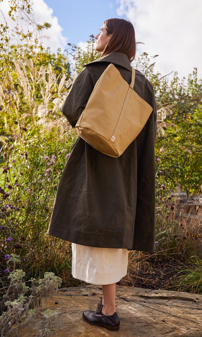 Plümo- Woman outdoors in a green coat stands on a rock with a canvas tool bag, looking up at the sky.