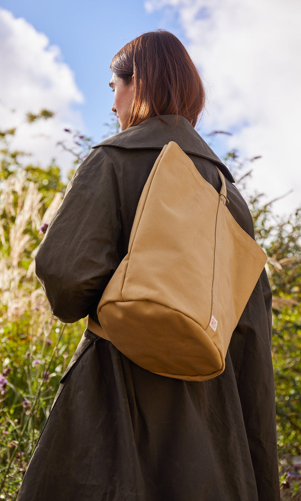 Plümo- Woman outdoors in an olive coat carries a Canvas tool bag; greenery and sky form the background.