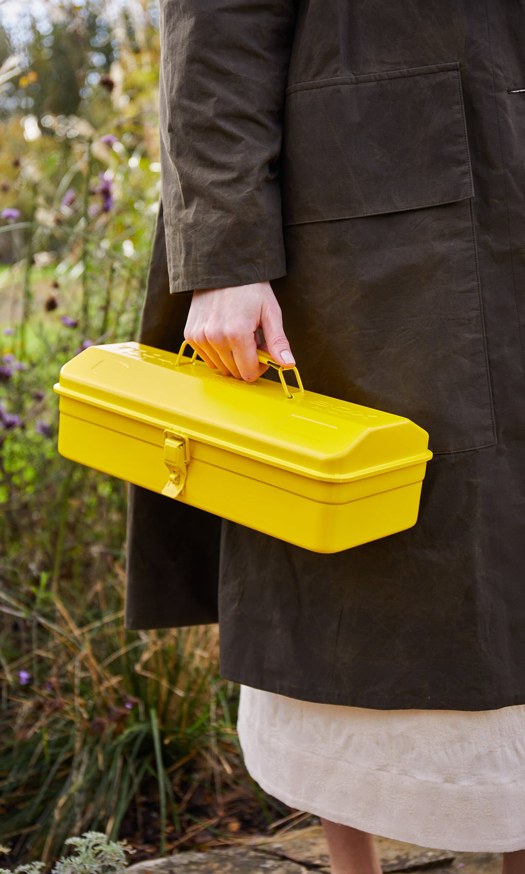 Plümo- A person in a dark coat holds a Yellow tool box near plants and flowers outdoors.