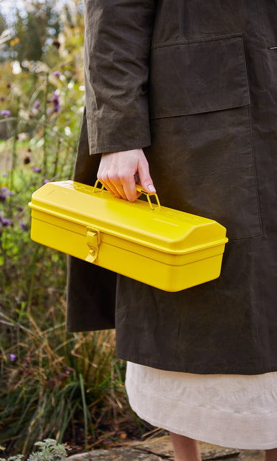 Plümo- A person in a dark coat holds a Yellow tool box near plants and flowers outdoors.