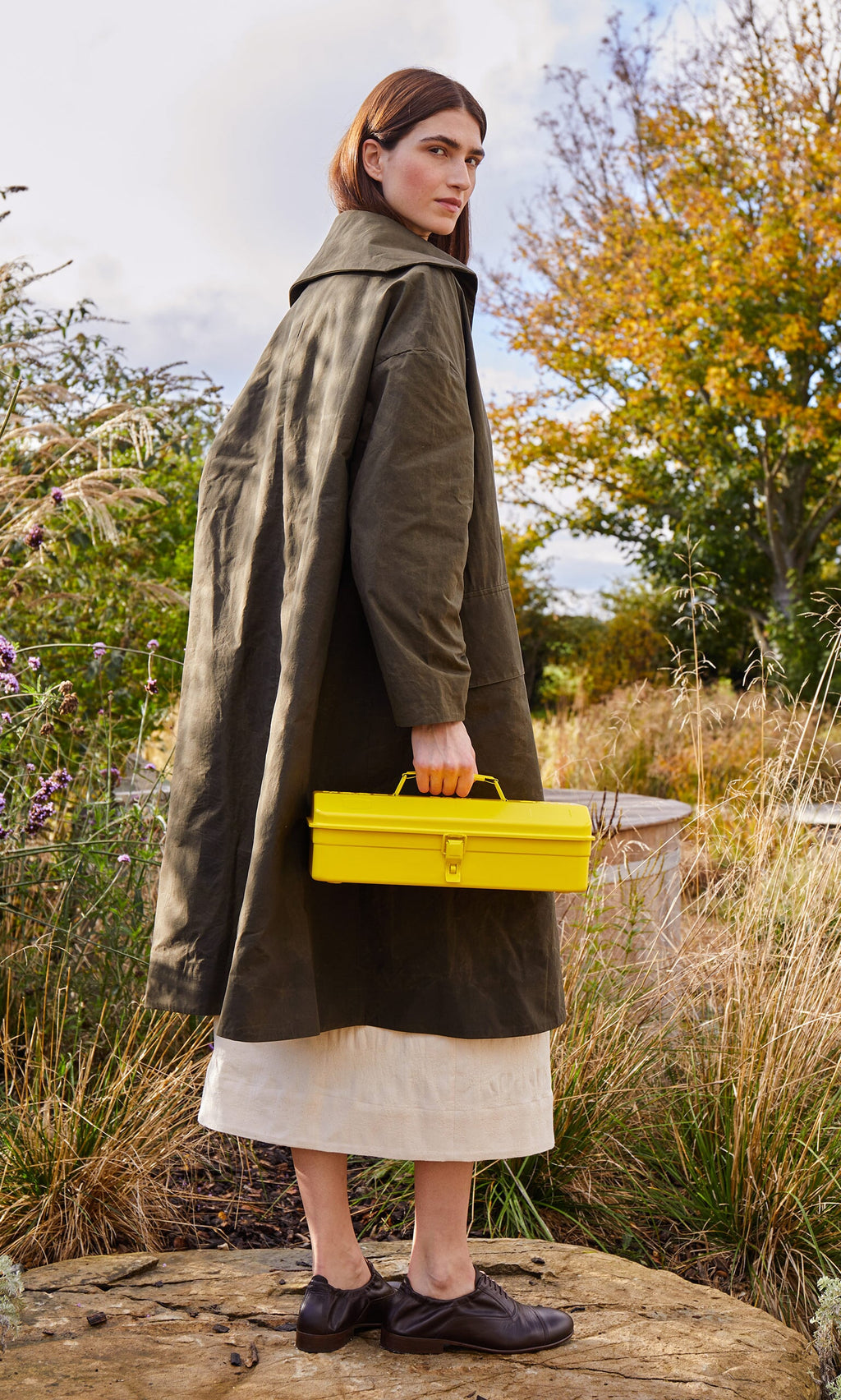 Plümo- A woman in an olive green coat holds a Yellow tool box with autumn trees in the background.