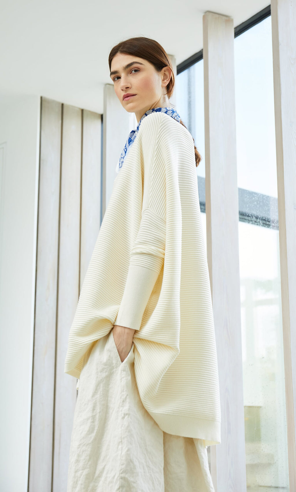Plümo- Woman in the Jack Sweater - Cloud stands indoors by tall windows, looking over her shoulder.