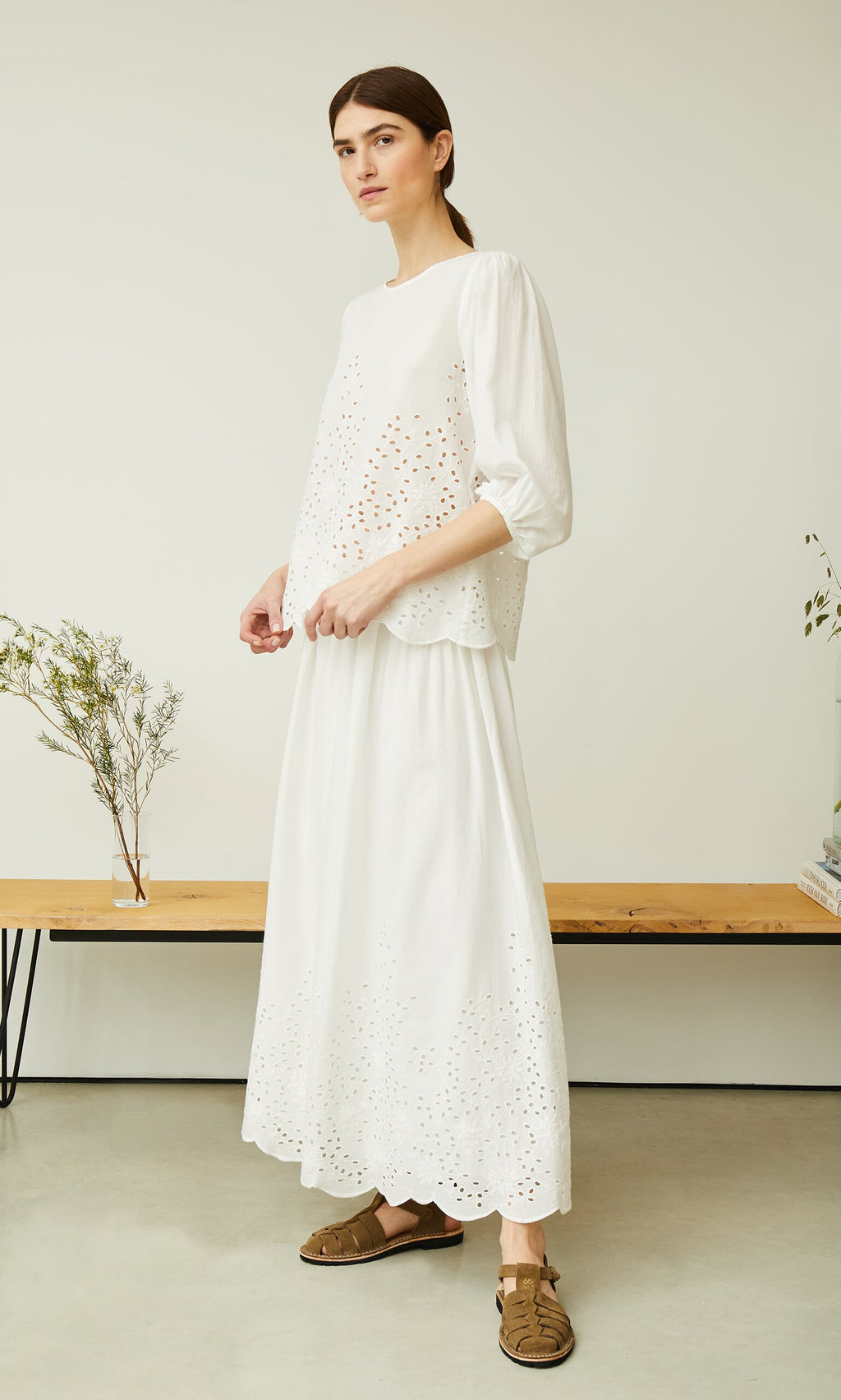 Plümo- Woman in a white broderie anglaise top, Bright Skirt, and brown sandals by a wooden bench with plants.