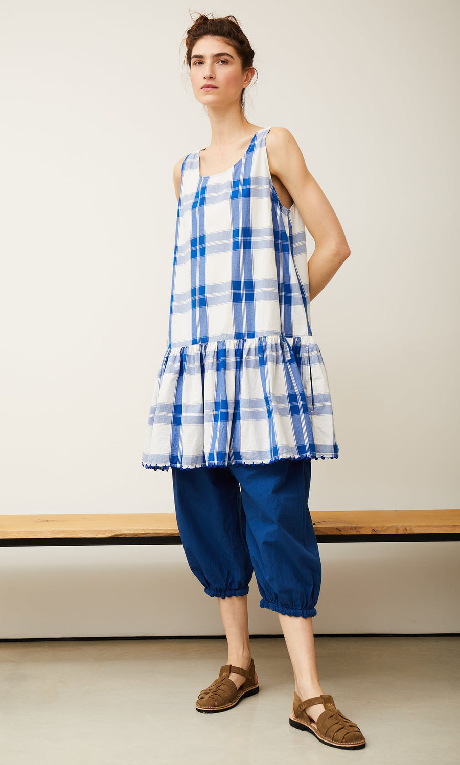 Plümo- Woman in a Swedish Swale top by Ewa i Walla over blue pants and brown sandals, standing indoors by a wooden bench.