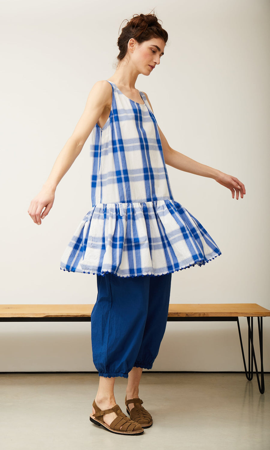 Plümo- Woman in a Swedish Swale top by Ewa i Walla and blue pants, arms outstretched, stands sideways in a minimalist room.
