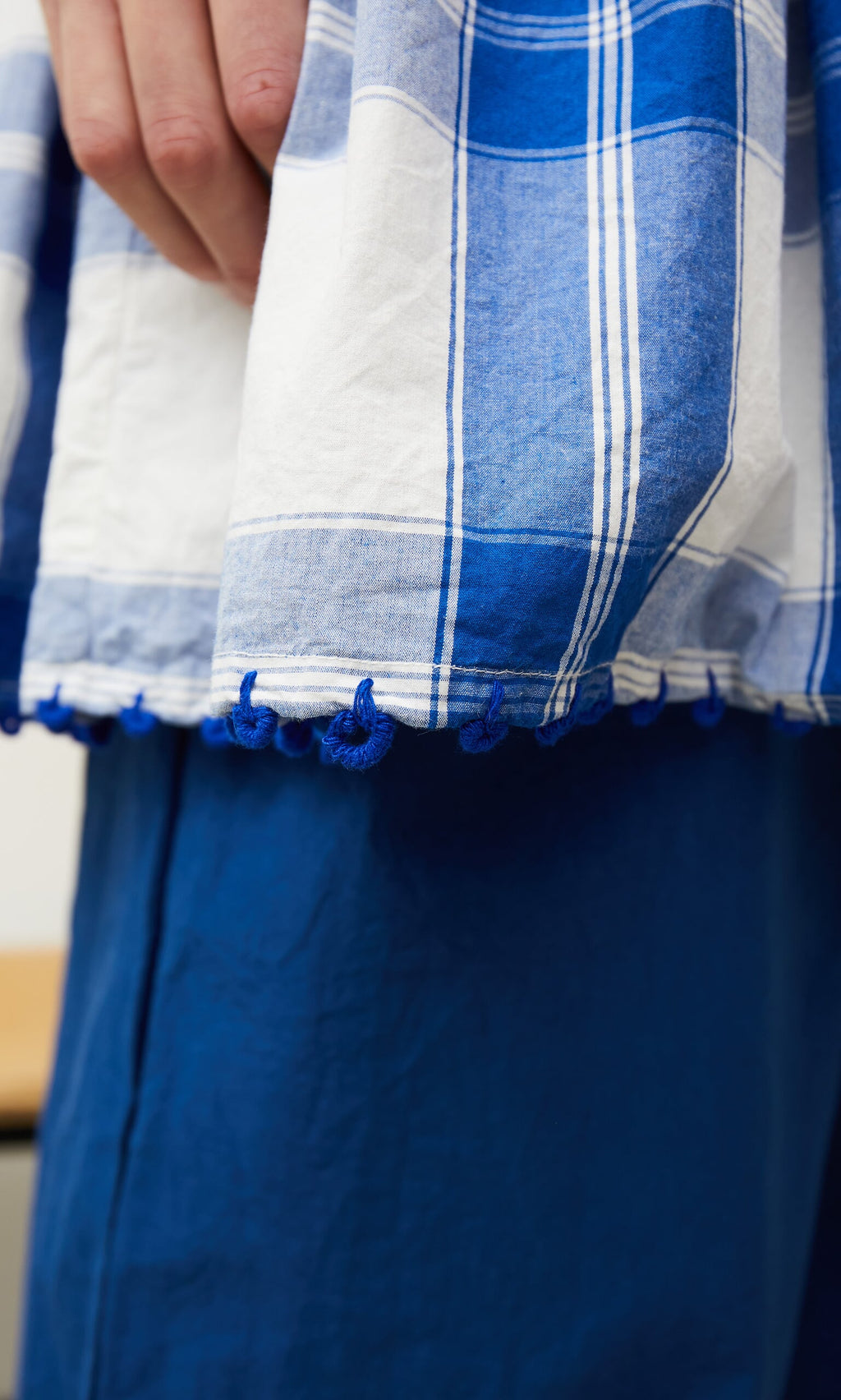 Plümo- Close-up of a hand holding the Swedish Swale top by Ewa i Walla, featuring blue and white checks over blue pants.