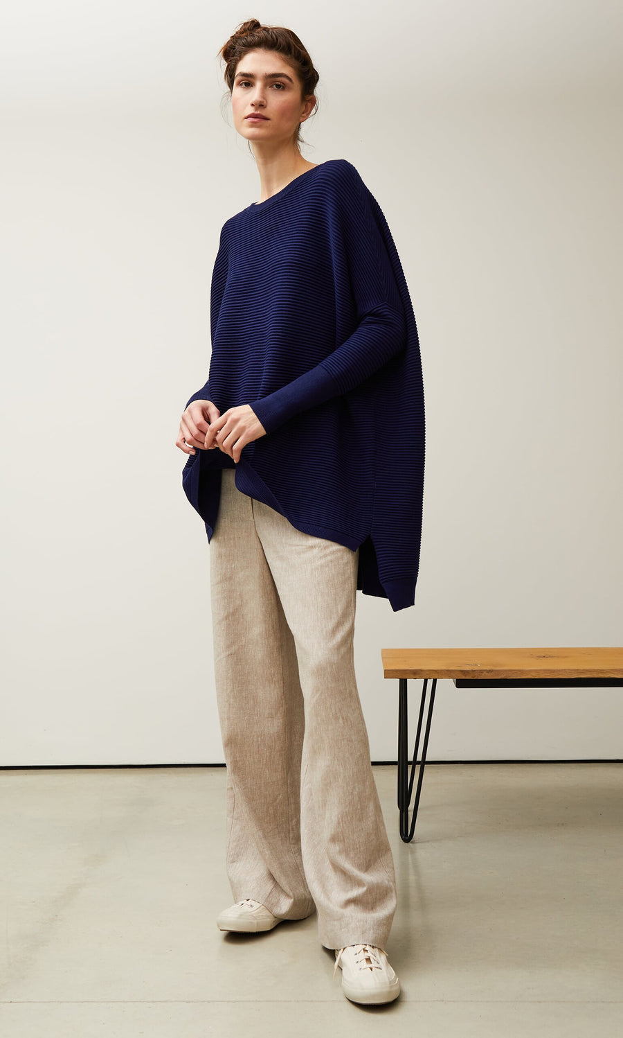 Plümo- Woman in the Jack Sweater - Navy, beige wide-leg pants, and white sneakers stands by a bench in a minimal room.