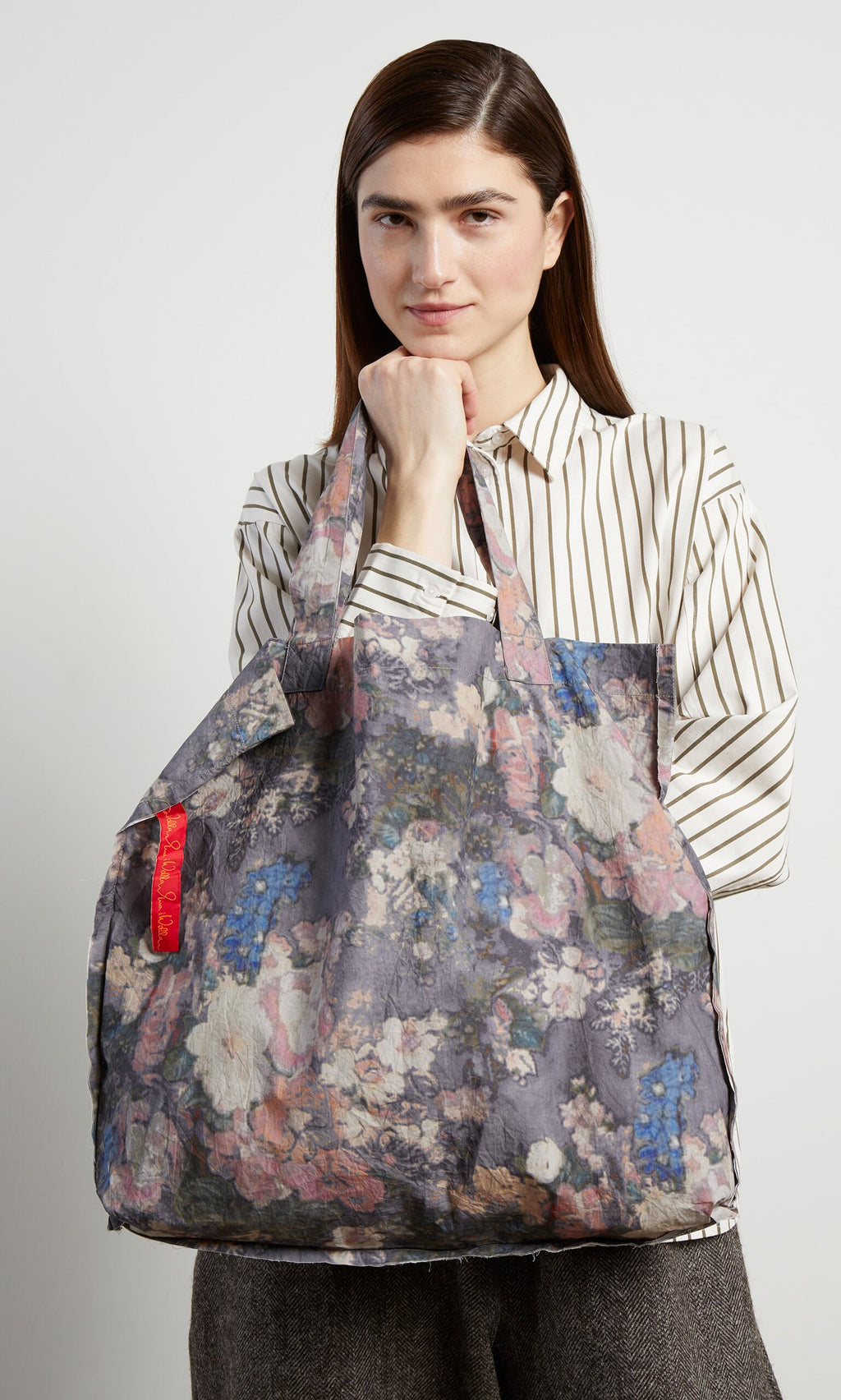 Plümo- Woman in a striped shirt holds the Manet Bag, a large floral print shopper with a red tag, against a plain background.