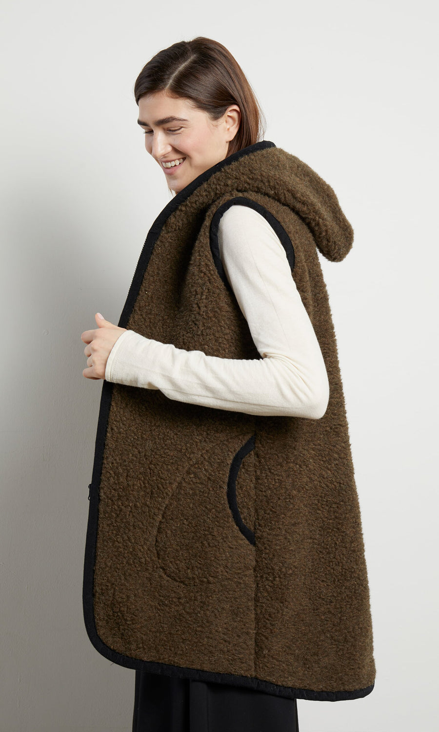 Plümo- A woman smiles sideways in the Bigger Rambler Gilet - Peat Brown, layered over a hooded fleece against a plain backdrop.