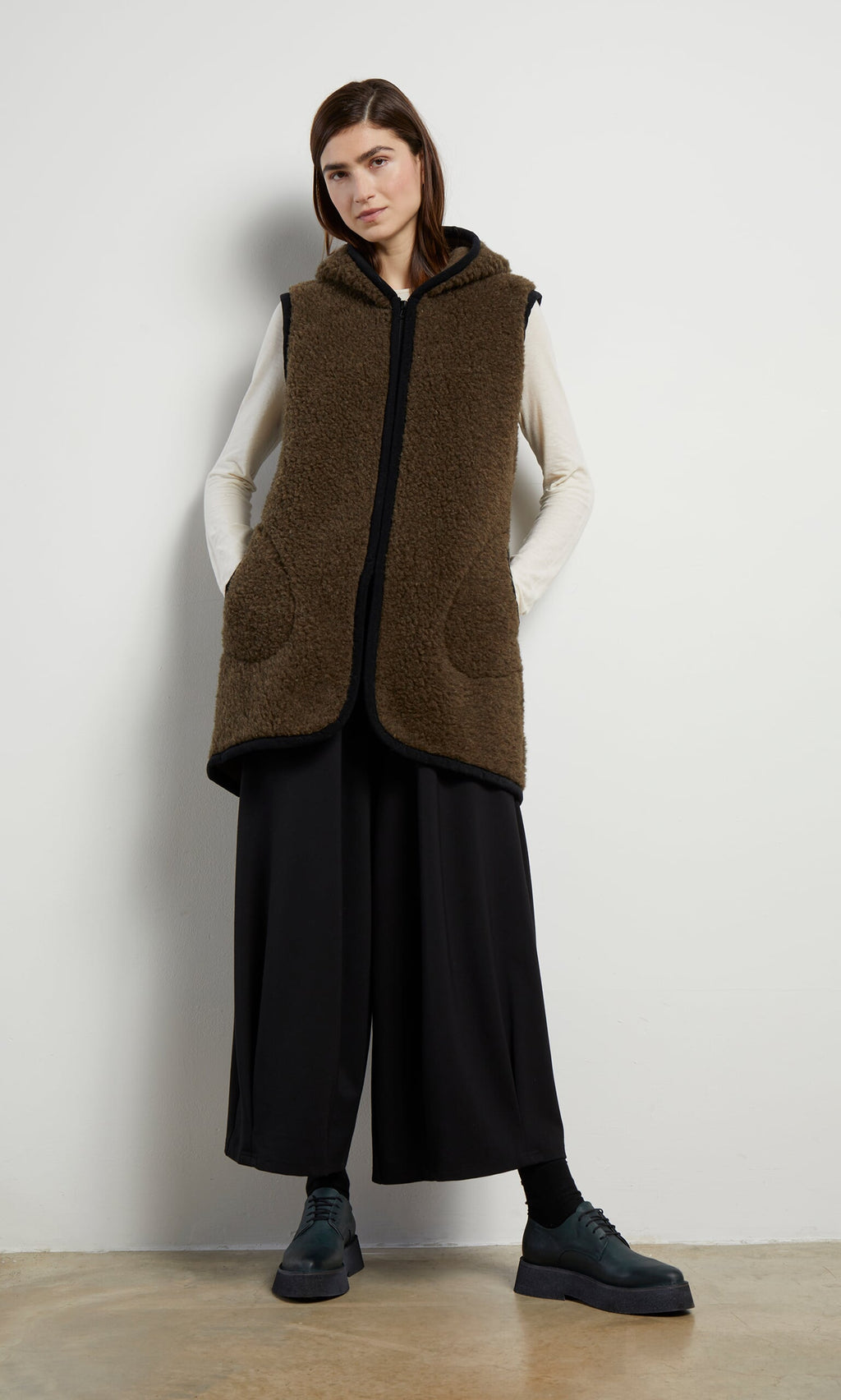 Plümo- Woman wearing the Bigger Rambler Gilet in Peat Brown, cream top, black pants, and black shoes, standing indoors.