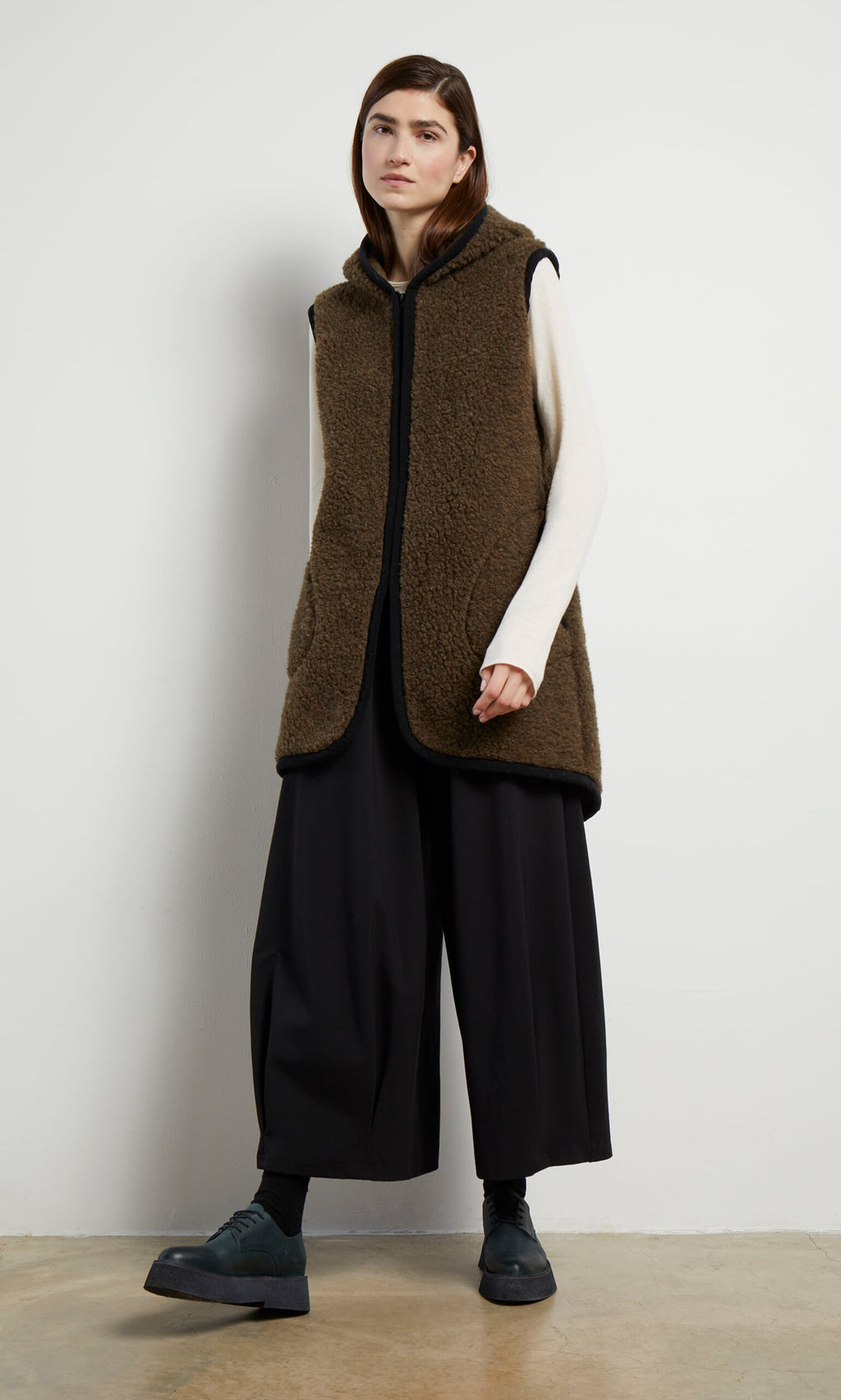 Plümo- Someone wears the Bigger Rambler Gilet in Peat Brown over a cream top, with black wide-leg pants and dark shoes indoors.