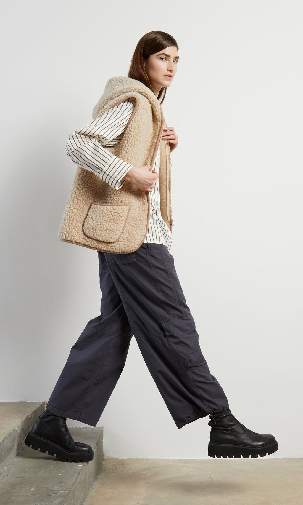 Plümo- Woman wearing the Wool Gilet - Natural, a striped shirt, and dark pants steps up stairs in black platform boots.