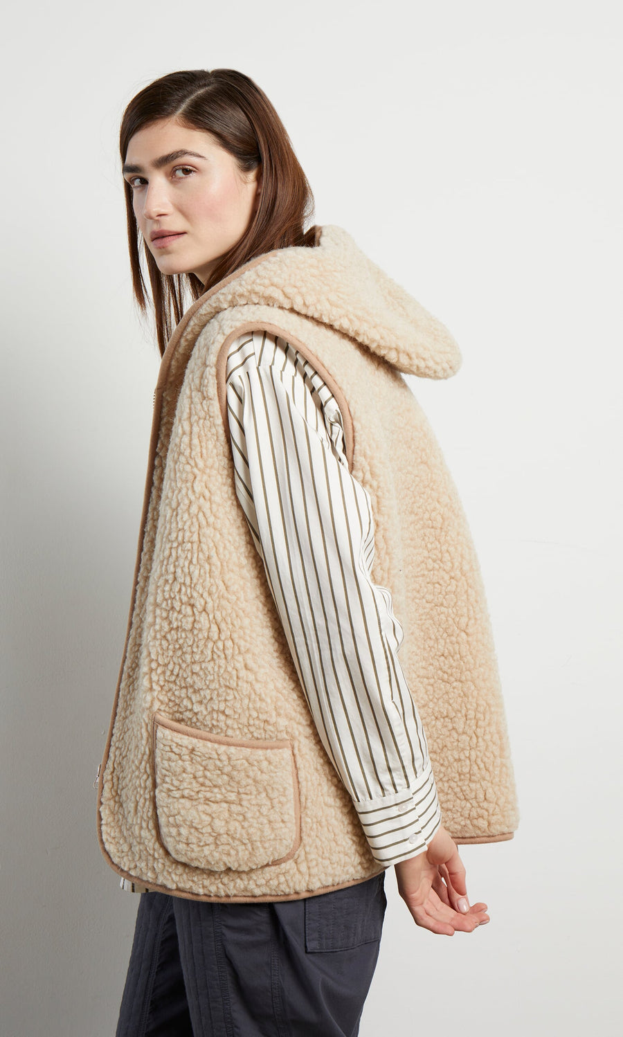 Plümo- Woman in a Wool Gilet - Natural, striped shirt, and dark pants, standing sideways against a white background.
