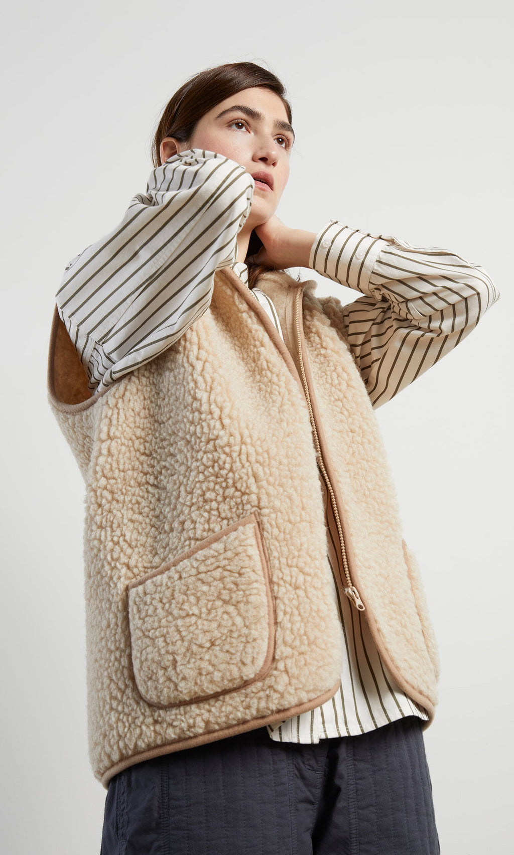 Plümo- A person in the Wool Gilet - Natural over a striped shirt adjusts the collar, looking slightly upward.